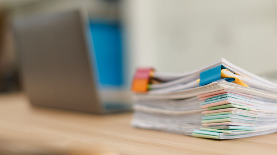 Stack of document files, paperwork, and reports organized with binder clips on a desk next to a blurred laptop