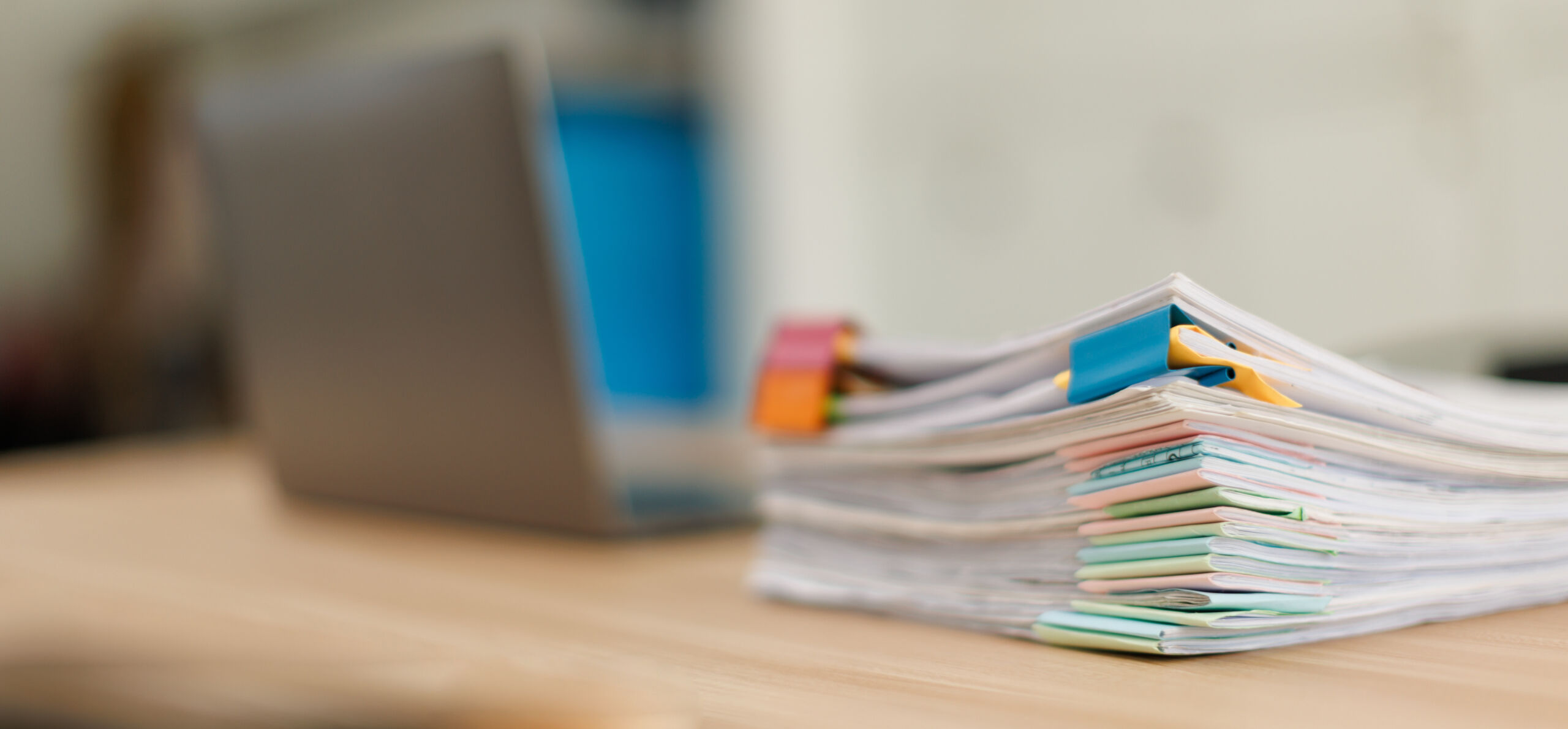 Stack of document files, paperwork, and reports organized with binder clips on a desk next to a blurred laptop