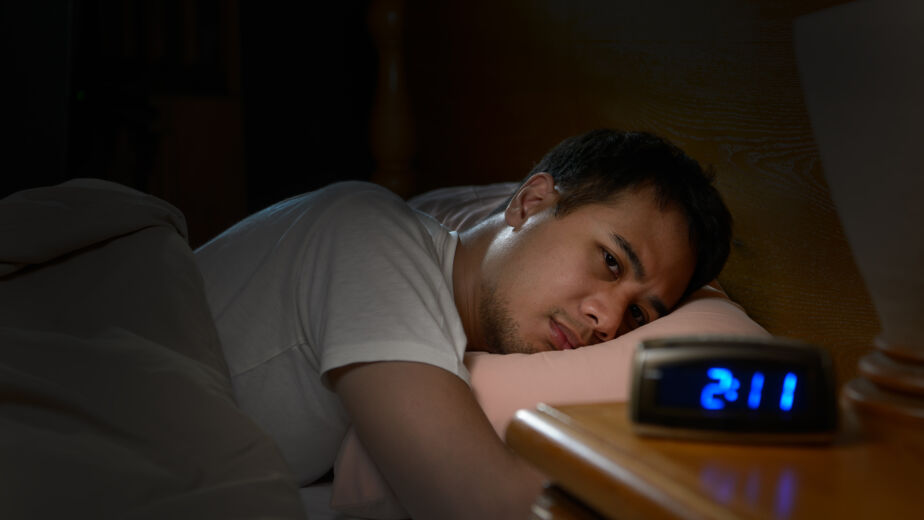 Mensch liegt schlaflos im Bett und schaut den Wecker an, der 2:11 in der Früh zeigt.